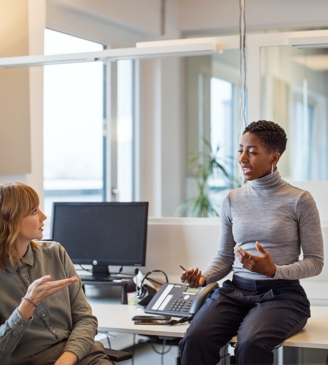 Black-woman-sitting-on-work-table-talking-to-white-woman-content-photo
